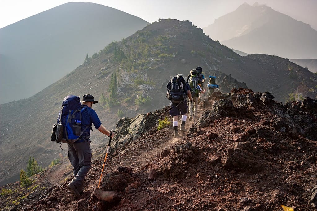PCT Trail Conditions That Hikers Need To Know A group of hikers with backpacks and trekking poles walk along a rocky mountain trail under a hazy sky.